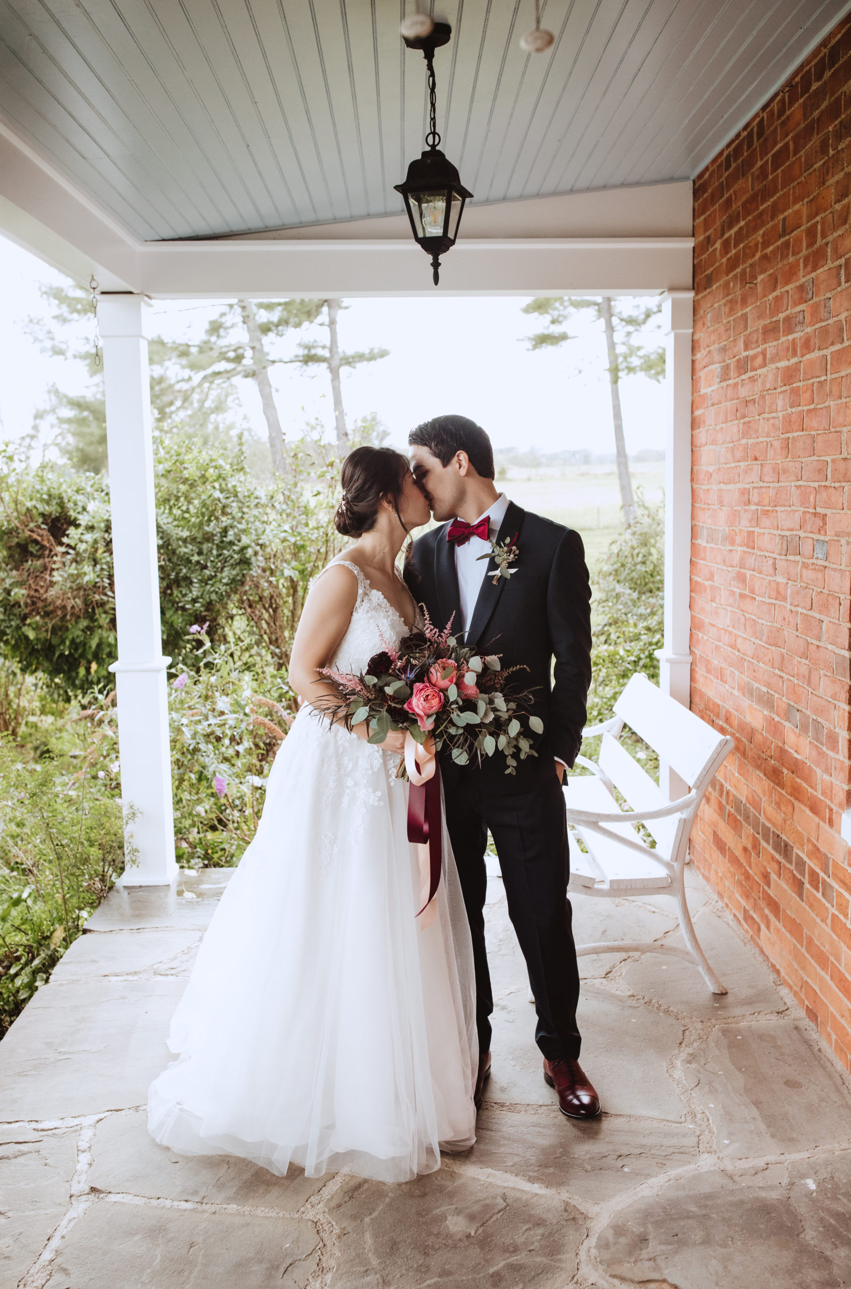 bride and groom kissing in the farmhouse porch. Bride is holding a garden style hand tie bouquet